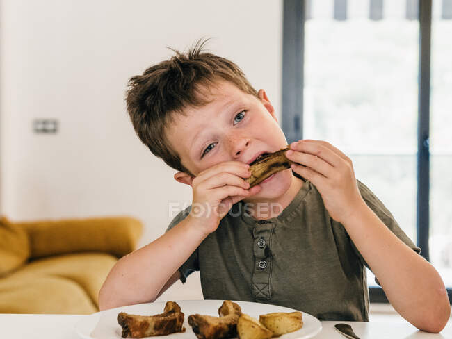 Очаровательный ребенок ест аппетитные свиные ребрышки во время обеда дома и смотрит в сторону — стоковое фото