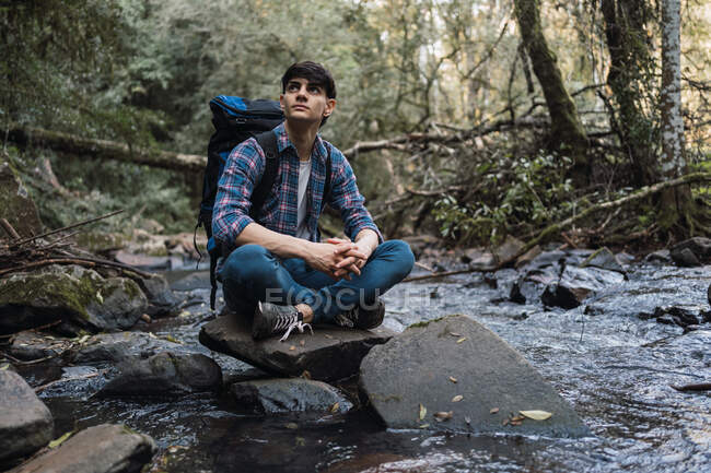 Side view of content male hiker with backpack sitting near lake in woods and browsing mobile phone while traveling — Stock Photo
