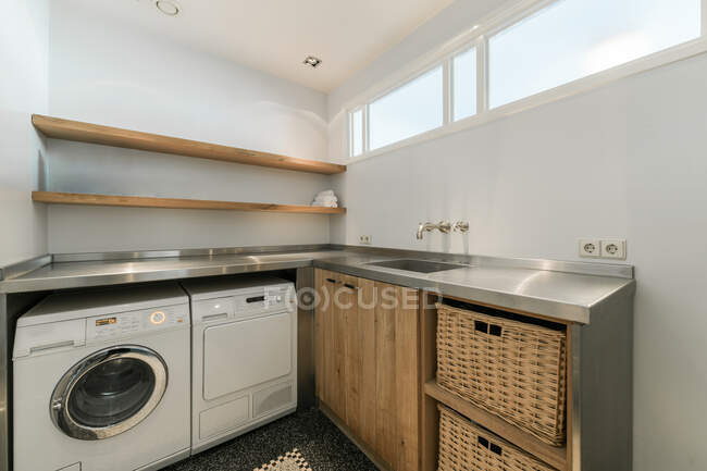 Innengestaltung der kleinen Waschküche mit Waschmaschine und Weidenkörben unter Metalltheke mit Waschbecken in moderner Wohnung — Stockfoto