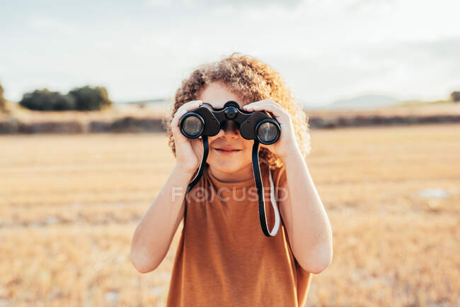 Delighted ethnic child with Afro hairstyle looking through binoculars and celebrating victory with raised arms while standing in dried filed in summer on sunny day and having fun — Stock Photo