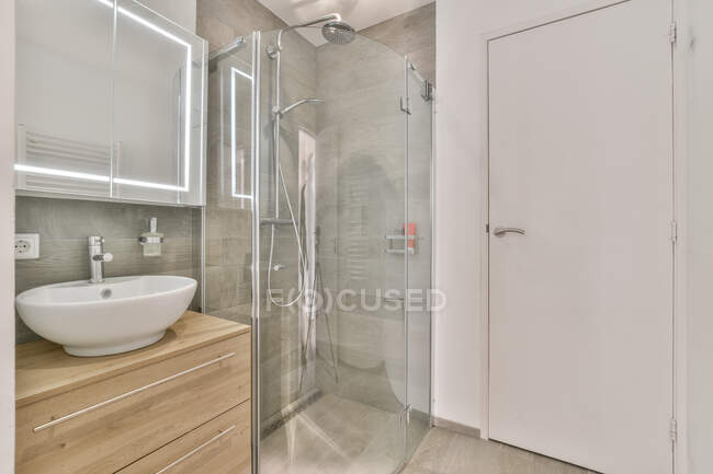 Stylish white bathroom interior with washbasin and wooden cabinet near shower cabin with glass partition in modern apartment — Stock Photo