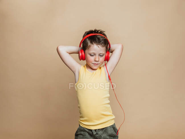 Menino pré-adolescente despreocupado em fones de ouvido vermelhos ouvindo música enquanto estava em pé no fundo marrom e olhando para a câmera — Fotografia de Stock