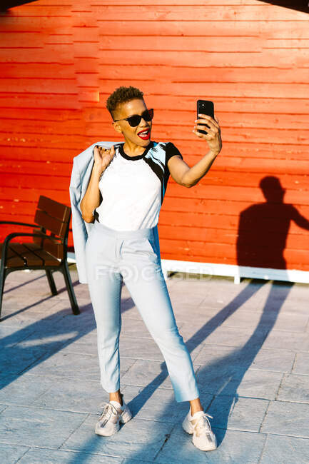 Allegro afro-americano donna in abito alla moda con mano in tasca parlando sul cellulare, mentre guardando altrove in città soleggiata — Foto stock