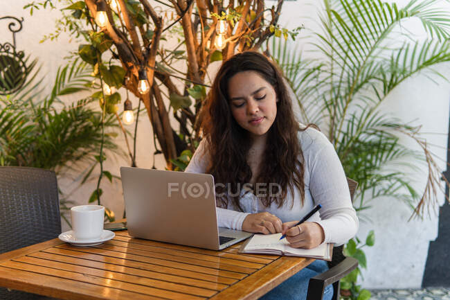 Joven estudiante latinoamericana enfocada en tomar notas en planificador mientras trabaja en la computadora portátil preparándose para el examen universitario en la acogedora cafetería - foto de stock