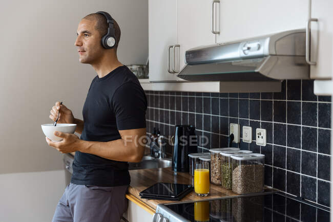 Чоловік в навушниках слухає музику і снідає зі свіжим апельсиновим соком вранці вдома і стоїть біля стійки на кухні, дивлячись далеко — стокове фото