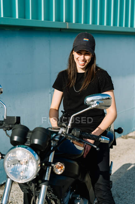 Motociclista feminina positiva sentada em moto estacionada na rua da cidade no dia ensolarado e olhando para a câmera — Fotografia de Stock