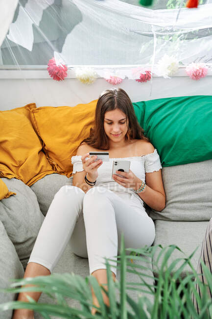 Jeune femme de contenu assis sur le canapé et effectuant le paiement avec une carte en plastique pour commander lors des achats en ligne sur téléphone mobile — Photo de stock