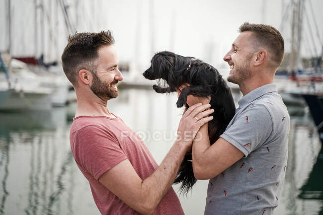 Side view of cheerful adult homosexual men with cute dog looking at each other in harbor — Stock Photo