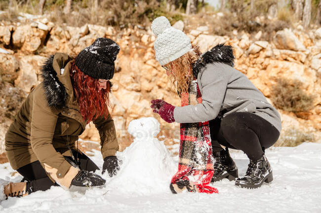 Vista lateral de amigas em outerwear fazendo boneco de neve na floresta e se divertindo juntos durante o fim de semana — Fotografia de Stock