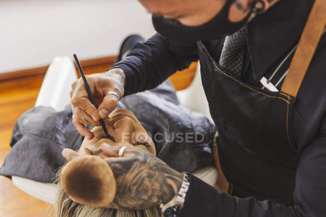Alto angolo di uomo tatuato in maschera disegno frecce eyeliner sulle palpebre della donna durante il lavoro in studio di trucco — Foto stock