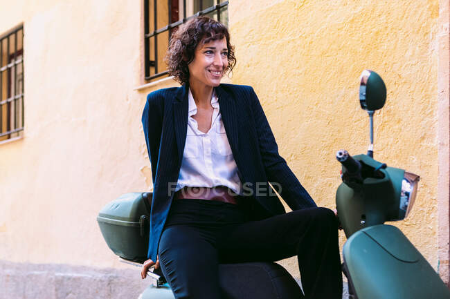 Positive female with curly hair wearing elegant outfit sitting on motorbike and looking away on street — Stock Photo