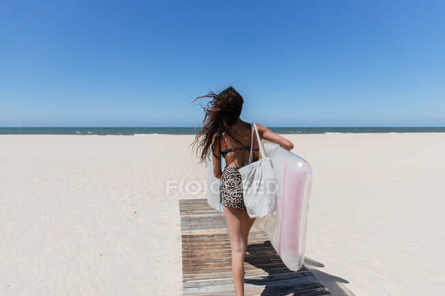 Back view of positive female in summer outfit and with inflatable mattress walking on sandy coast on sunny day during vacation — Stock Photo
