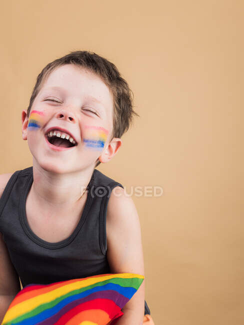 Fröhliches Kind mit geschminkten Wangen und LGBTQ-Flagge auf beigem Hintergrund — Stockfoto