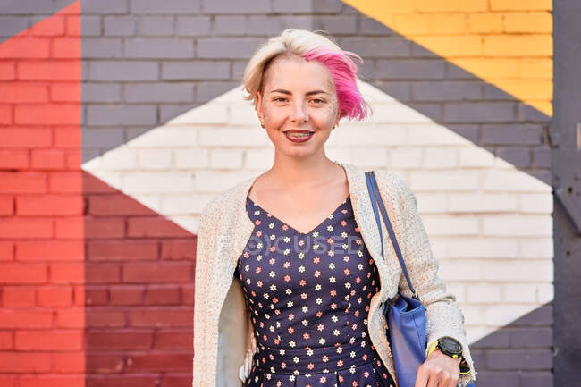 Happy fêmea alternativa no vestido da moda e com cabelo curto tingido de pé contra a parede de tijolo colorido na rua e olhando para a câmera — Fotografia de Stock