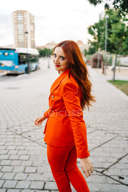 Vista posteriore di donna sicura con lunghi capelli rossicci e in abito rosso vivido alla moda che cammina in città e guarda la fotocamera — Foto stock