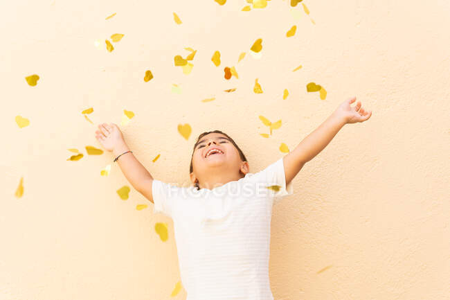 Joyeux garçon en t-shirt blanc debout avec les bras levés tout en jetant pile de confettis d'ouïe jaune sur le mur orange clair — Photo de stock