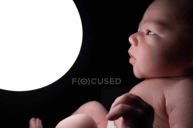 Side view of tender infant on bed and touching glowing night light lamp in dark room — Stock Photo
