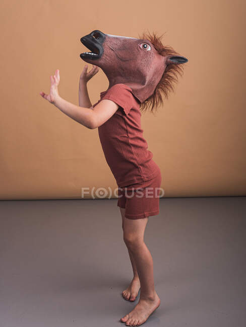 Vista lateral del niño anónimo con máscara de caballo inclinada hacia adelante mientras representa el concepto de semental galopante sobre fondo beige - foto de stock