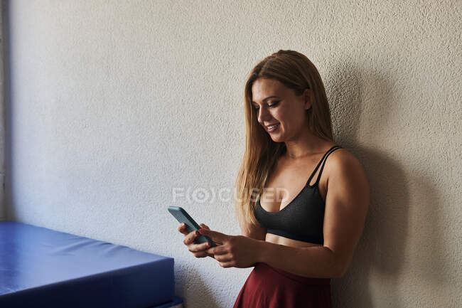 Веселая спортсменка стоит рядом с ковриком в спортзале и просматривает мобильный телефон — стоковое фото