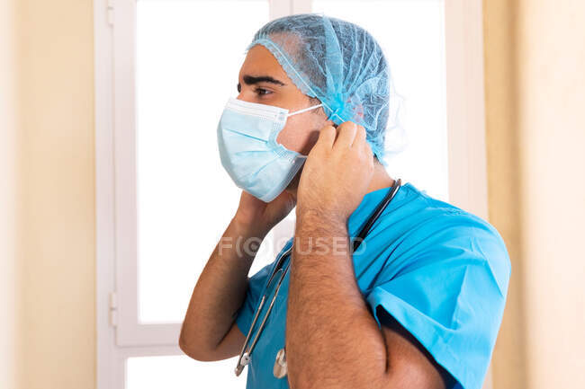 Side view of male medic in uniform putting on disposable mask during work in clinic — Stock Photo