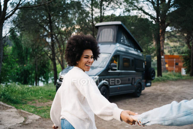 Smiling African American woman holding hands of cropped boyfriend holding hands while strolling and looking away against camper — Stock Photo
