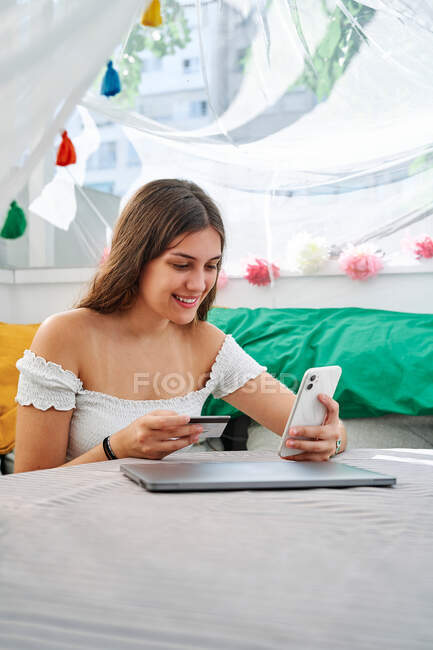 Молодий контент жінка сидить на столі і робить оплату пластиковою карткою для замовлення під час онлайн-покупки на мобільний телефон — стокове фото