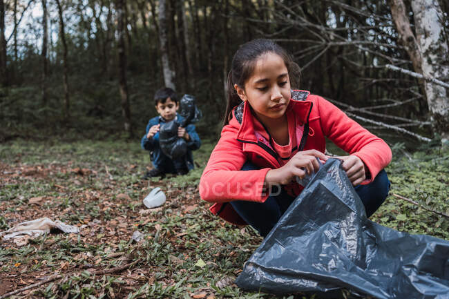 Ethnic volunteers with plastic bags picking rubbish from terrain against trees in summer woods in daylight — Stock Photo