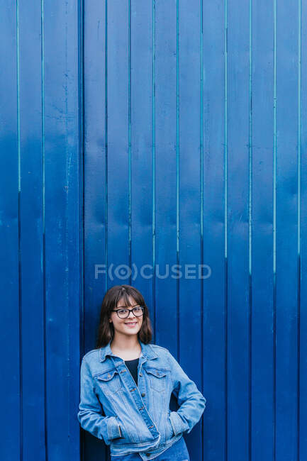 Trendy female hipster wearing denim jacket and jeans leaning on blue wall in city street and looking at camera — Stock Photo