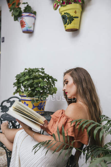Seitenansicht der anmutigen Frau im Rock lehnt Beine auf Stuhl beim Chillen im Innenhof im Sommer und genießen interessante Geschichte in Buch am Wochenende — Stockfoto