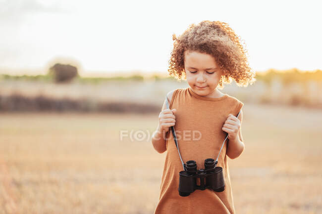Nettes ethnisches Kind mit lockigem Haar und Fernglas, das im Sommer auf einem getrockneten Feld steht und nach unten schaut — Stockfoto