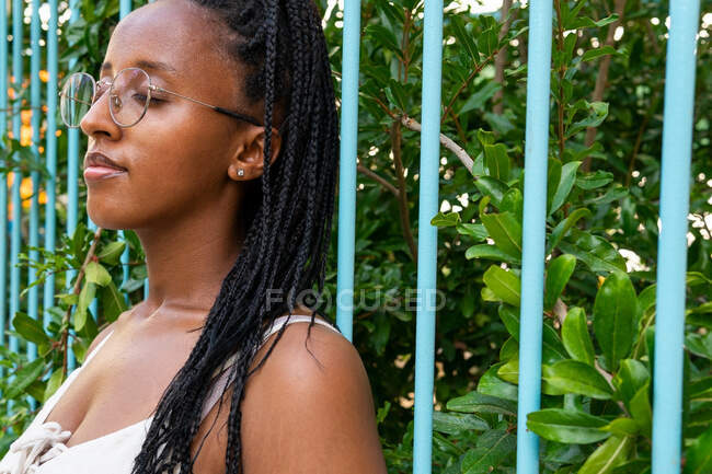 Tranquillo femmina afroamericana con lunghi capelli intrecciati in piedi vicino alla recinzione in giardino verde in estate e godersi la natura con gli occhi chiusi a Barcellona — Foto stock