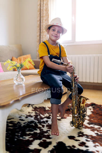 Cheerful kid in hat with saxophone looking at camera while sitting on table in house room — Stock Photo