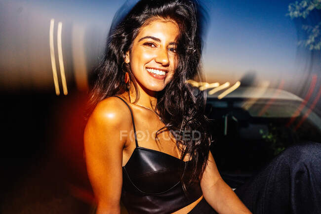 Attractive happy young long haired Hispanic brunette in black top with bare shoulders looking at camera sitting in a car with light reflection at night time — Stock Photo