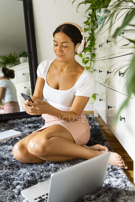 Cheerful ethnic female with cellphone listening to song from wireless headphones while sitting on carpet against mirror at home — Stock Photo