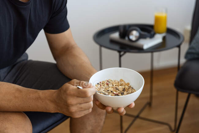 Macho de cultura anônimo comendo muesli saboroso da tigela enquanto sentado na cadeira em casa e tomando café da manhã saudável de manhã — Fotografia de Stock