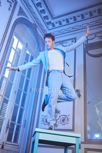 Von unten balanciert ein ausdrucksstarker Mann in stylischem Anzug auf einem Tisch im noblen Zimmer mit blauem Neonlicht und blickt weg — Stockfoto