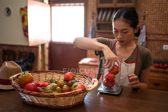Femme au foyer ethnique concentrée pesant des tomates fraîches dans une cruche en verre sur une balance de cuisine pendant la cuisson des aliments à la maison — Photo de stock