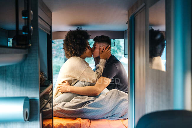 Vista lateral do jovem casal multirracial apaixonado beijando uns aos outros enquanto sentado na cama dentro van campista estacionado na natureza durante as férias românticas juntos no verão — Fotografia de Stock