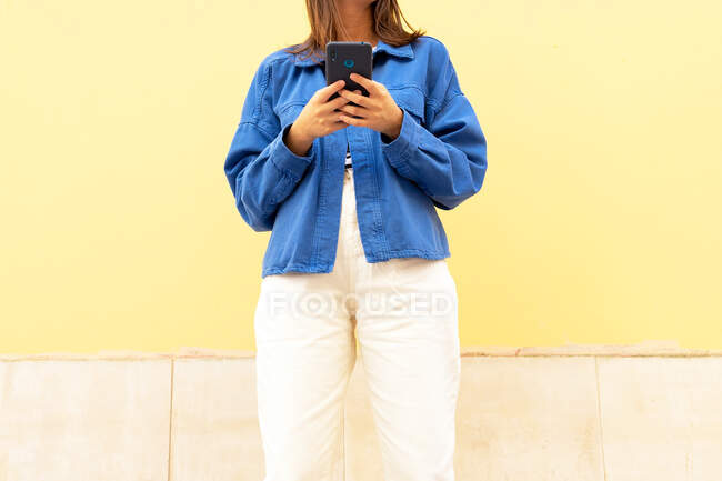 Recortado irreconocible joven mujer en traje de moda de mensajería en el teléfono móvil en el fondo de la pared en la calle de la ciudad y mirando hacia otro lado - foto de stock