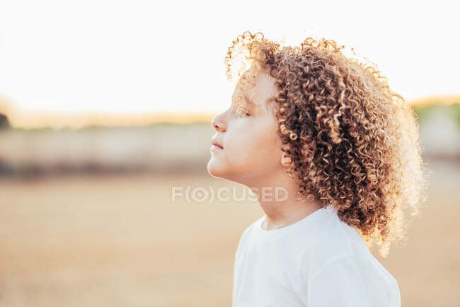 Vista lateral de adorable niño étnico con peinado afro y en camiseta blanca mirando hacia otro lado en el campo seco en verano en la espalda iluminada - foto de stock