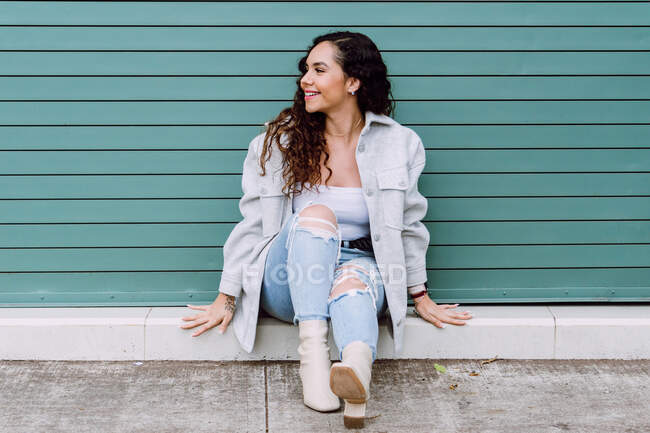 Charmante Frau in stilvoller Kleidung sitzt in der Nähe der Hauswand, während sie lächelt und wegschaut — Stockfoto