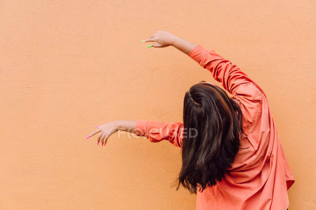 Back view of young anonymous woman in modern outfit with bright long manicure raising hands against orange background — Stock Photo
