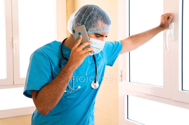 Чоловічий лікар у формі та зі стетоскопом, що стоїть у лікарні та розмовляє на смартфоні — стокове фото