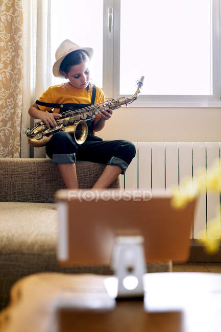 Ребенок играет на саксофоне, сидя на диване, записывая видео на мобильный телефон у окна в комнате — стоковое фото