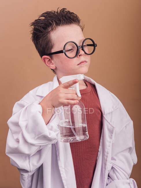 Enfant chimiste en verres décoratifs et peignoir de laboratoire regardant loin avec de l'eau dans le flacon distributeur sur fond beige — Photo de stock