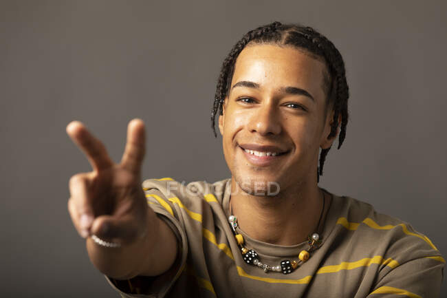 Heureux jeune afro-américain hipster gars avec des tresses portant chemise rayée et collier regardant la caméra et montrant deux doigts signe sur fond gris — Photo de stock