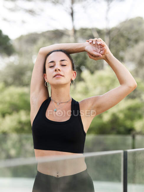Молодая спортсменка делает упражнения на растяжку возле стеклянных перил во время тренировки летом — стоковое фото
