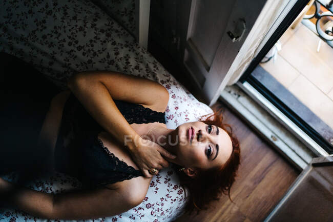 Top view of unemotional pretty redhead female lying on soft bed looking at camera at home — Stock Photo