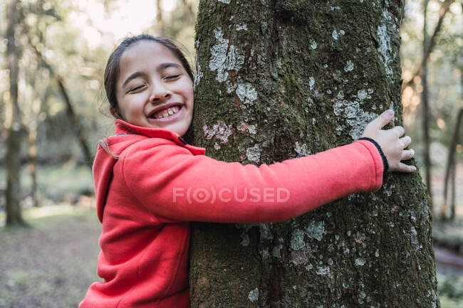 Vista lateral del encantador niño étnico tocando la corteza áspera del tronco de árbol envejecido con liquen con los ojos cerrados en el bosque - foto de stock