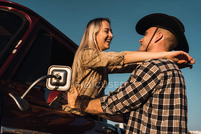 Vista lateral da mulher encantada saindo da janela do carro vintage e abraçando o namorado na camisa xadrez e chapéu de cowboy ao pôr do sol no campo — Fotografia de Stock